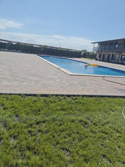 The image depicts an outdoor swimming pool area on a sunny day in Shkurupiivka, Ukraine. The pool, filled with clear blue water, is surrounded by a paved patio made of interlocking bricks in shades of brown and gray. In the pool, a yellow inflatable ring and a red and white ball are visible.
To the left of the pool, a dark green hedge forms a boundary, behind which rows of lounge chairs are arranged. Further in the background, trees and a small building with a wooden roof are visible.
To the right of the pool, a two-story building with balconies, windows, and air conditioning units is situated. The building appears to be a hotel or guesthouse, with multiple rooms visible on both floors. The sky above is a clear blue with scattered white clouds. In the foreground, a patch of green grass is present. A green garden hose is visible near the edge of the pool.