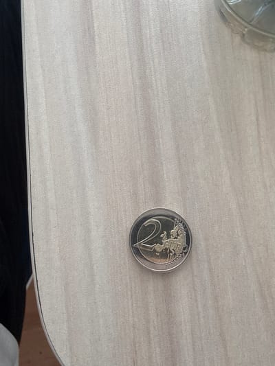A single two-euro coin lies on a light-colored, wood-grain textured surface. The coin is silver and gold, with the number "2" prominently displayed on the left side. The right side of the coin shows a depiction of the European continent. The coin is slightly angled. The background is mostly out of focus, with a dark, blurry area on the left and a partially visible glass object at the top right. The scene is devoid of any human presence or activity. The location is not discernible from the visual information provided.