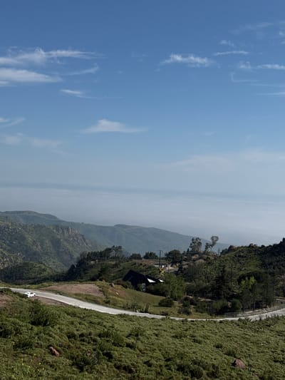 An elevated, wide-angle photograph depicts a mountainous landscape in the Unincorporated Santa Monica Mountains, United States of America, under clear daylight conditions. The sky occupies the upper third of the frame, displaying a light blue hue with sparse, wispy white clouds. The horizon line, marked by atmospheric haze, reveals distant mountains and what appears to be the Pacific Ocean.
In the mid-ground, a paved two-lane road, marked with white lines, curves from the lower-left to the center-right. A single white passenger vehicle, appearing to be a sedan or compact SUV, is visible on the road in the mid-left, oriented towards the right. To the right of the road, a dark-colored building with a sloped, angular roof is situated on a hillside, partially obscured by trees.
The terrain is characterized by rolling hills and steep slopes covered predominantly in green, low-lying brush and scattered trees. Exposed reddish-brown rocks are visible throughout the landscape, particularly on the lower left and in the vicinity of the building. Several utility poles with power lines traverse the hillsides. No individuals are visible in the scene.