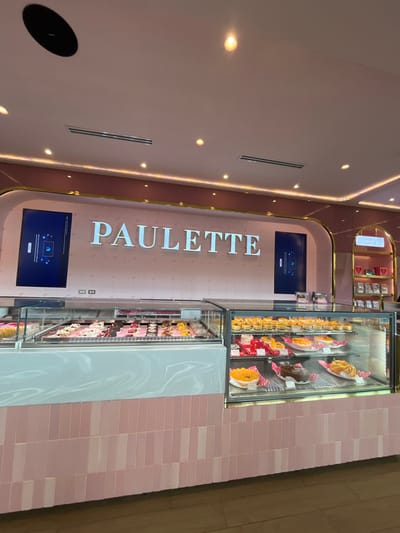 The image shows a sweets shop called "Paulette". The store has a sleek, modern design with pink walls and gold accents. A display case is filled with a variety of pastries and cakes. There is a digital screen at the top and bottom. The wall to the right of the display case is covered in pink tiles.