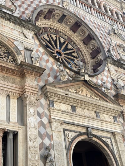 This is a close-up, low-angle shot of the facade of a building, likely a cathedral or a historical monument, characterized by intricate architectural details. The building features a repeating diamond pattern in shades of pinkish-brown, cream, and grey, creating a visually striking mosaic effect. Above this pattern, a large, ornate rose window is visible, its intricate stone tracery forming a radial design. The window appears to be made of dark wood or metal spokes against a darker background, possibly glass.
Below the rose window, the facade is adorned with multiple layers of carved stone. These carvings include decorative moldings, friezes with figural reliefs, and classical architectural elements like columns and pediments. Several small statues of figures, likely religious in nature, are placed on ledges and at architectural junctures. One statue on the lower left appears to be a winged figure. Another, on the lower right, is a standing female figure. The architecture suggests a Gothic or Renaissance style.
The lighting suggests it is daytime, with soft light illuminating the details without harsh shadows, possibly indicating an overcast day or late afternoon light. The stone surfaces show signs of age and weathering, contributing to the historical feel of the structure. The scene is static, focusing entirely on the architectural grandeur of the building. No people are clearly identifiable as active participants in a scene, only the sculpted figures. The location context points to Mozzo, Italy. No visible text is discernible in the image.