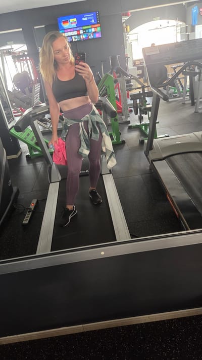 A woman is taking a selfie in a gym. She is wearing a black sports bra, purple leggings, and black sneakers. A green and white plaid shirt is tied around her waist. She is standing on a treadmill. In the background, there are various gym equipment, including weight machines and other treadmills. A television screen is mounted on the wall, displaying a menu with flags of Spain and Germany. The lighting suggests it is daytime. The woman appears relaxed and is looking at her phone, which she is using to take the picture. The gym is well-lit with modern equipment and a dark, speckled floor.