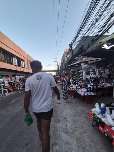 A man in a white t-shirt with a QR code on the back and dark shorts is walking down a street in Parañaque, Philippines. He is holding a green cloth in his right hand. Along the street, vendors have set up stands selling clothing and shoes. Power lines are strung overhead. Other people are visible, including one woman wearing a head covering and floral pants. A multistory building is on the left side of the street.