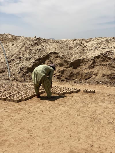 In Deh-E Bagh, Afghanistan, a man dressed in a light green shalwar kameez and a small patterned cap bends over a rectangular field of drying bricks. The bricks, arranged in neat rows, have a square indentation on top. Behind the man and the bricks is a large dirt embankment. A single pipe snakes along the top edge of the embankment. The ground is dry and dusty. The sky is pale blue.