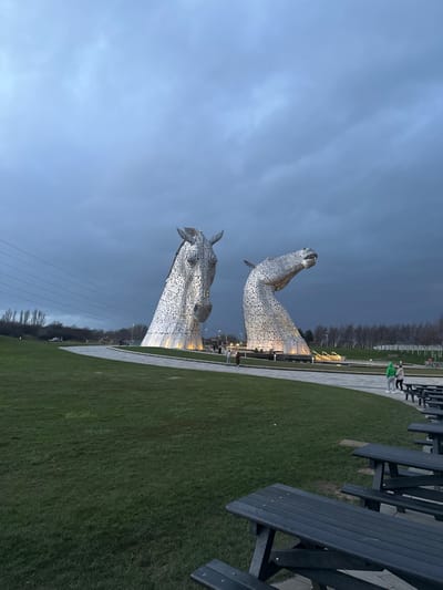 The image displays the Kelpies, two large, illuminated metallic horse-head sculptures, situated within Helix Park in Falkirk, United Kingdom, under a dark, overcast sky. The left sculpture, with its head slightly lowered, faces forward, while the right sculpture's head is tilted upwards and to the right. Both sculptures exhibit intricate construction from numerous overlapping metal plates and are illuminated from below by white lights.
In the midground, paved grey pathways curve around the bases of the sculptures. Several individuals are visible on these paths; a group of three is near the right sculpture, one wearing a distinctive light green/yellow hooded top, and two others are further back near the left sculpture. These individuals appear to be walking and observing the structures.
The foreground consists of a wide expanse of green grass. In the lower right corner, a row of dark grey wooden picnic tables with attached benches is partially visible, extending out of frame. The background features bare deciduous trees and distant industrial or urban infrastructure. Power lines run horizontally across the left side of the upper frame. The overall lighting suggests dusk or early evening conditions.