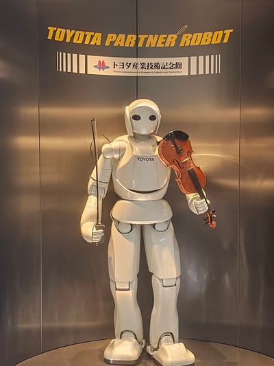 The image depicts a full-body view of a white, humanoid robot standing upright, holding a violin and a bow. The robot has a smooth, featureless head with two dark eye-like sensors. It is dressed in a white, segmented body suit, with visible joints at the shoulders, elbows, waist, knees, and ankles. The violin is positioned across the robot's chest, held by its right hand, while the bow is held vertically in its left hand, near the robot's side.

Above the robot, a large sign with yellow text reads "TOYOTA PARTNER ROBOT". Below this, a smaller sign with Japanese text and the Toyota logo reads "トヨタ産業技術記念館" (Toyota Commemorative Museum of Industry and Technology). The background is a metallic, reflective surface that appears to be part of an indoor exhibition space. The lighting is somewhat warm, casting reflections on the robot and the background.

This robot is located at the Toyota Commemorative Museum of Industry and Technology in Nagoya, Japan. The museum is dedicated to showcasing the history of the Toyota Group's manufacturing and technological innovations.