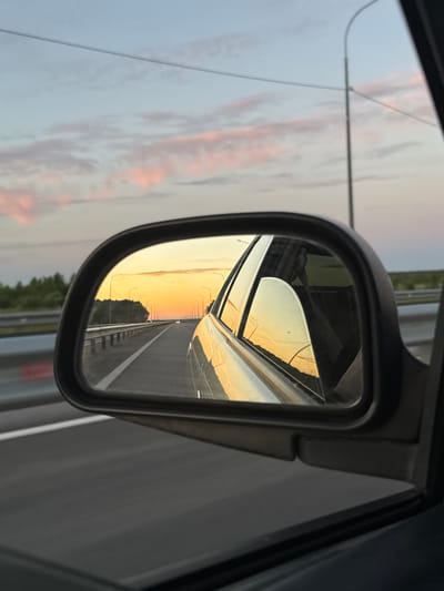 The image is a reflection in a car's side mirror, showing a highway at sunset. The sky is filled with soft orange and pink clouds. Streetlights line the highway, which appears to be in a rural or suburban area. The reflection also shows part of the car's interior, including the steering wheel and dashboard. The location is stated as Afanasovo, Russia.