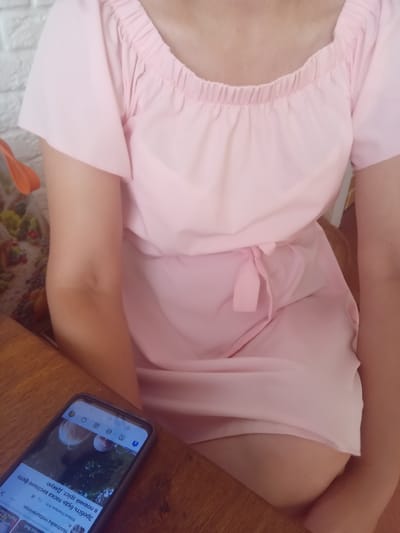 A person wearing a pink dress is seated at a wooden table with a smartphone in front of them. The dress has short, flared sleeves and an elasticized neckline. The smartphone displays social media content, including text in Cyrillic. The background includes a white brick wall and a bag with a floral pattern. The scene appears to be indoors, possibly in a cafe or restaurant in Terebovlia, Ukraine, given the Cyrillic text on the phone.