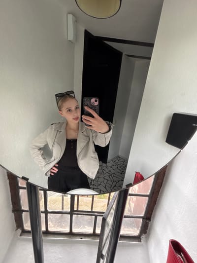 A young Caucasian female is depicted in a mirror selfie taken indoors in Hochheim, Germany. She has light-colored hair styled back, with dark-framed sunglasses resting on top of her head. She wears a light grey or off-white biker-style jacket over a black scoop-neck top and black trousers. A silver cross pendant on a thin chain and silver hoop earrings are visible. Her left hand is placed on her hip, and her right hand holds a smartphone with a dark, glittery case featuring a multi-lens camera module, positioned to capture her reflection. Her expression is playful, with her tongue slightly extended.

The mirror has an irregular, non-rectangular shape. Reflected in the mirror behind the subject's head is a white wall with a white rectangular device mounted high up, likely a sensor or thermostat. To the left, a dark doorway or recessed area is visible, with hints of a white ceiling above and a circular light fixture.

To the right of the subject's reflection, a white wall displays a black rectangular object mounted, possibly a speaker or security camera, with the partial letters "KE" visible on its surface. Below the mirror, a multi-pane window with dark frames is reflected, revealing an outdoor scene with diffused light, ground-level surfaces, and distant structures. A prominent dark vertical bar, possibly part of the window's structure or an adjacent railing, is also reflected. A small portion of a red object, resembling a bag or purse, is visible at the lower right edge of the mirror's reflection. The floor visible in the reflection to the lower left of the subject features an irregular black and white mosaic tile pattern.