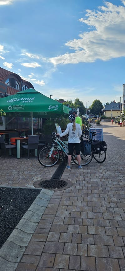 Two people, a woman and a man, are standing next to their bicycles on a paved area with a patterned brick surface. The woman, wearing a white long-sleeved shirt and black shorts, is holding a map or paper. The man, in a bright green shirt and helmet, is also looking at the paper. Both are wearing helmets, suggesting they are cyclists. Their bicycles are equipped with panniers, indicating they might be on a tour or journey.
Behind them, a green umbrella with the "Soproni 0.0%" logo is providing shade over an outdoor seating area with tables and chairs. The umbrella also displays text about the product being made from recycled plastic. A sign board with the word "ANGEBOT" (German for "offer") is visible behind the bicycles.
The scene is set in an outdoor environment with buildings and trees visible in the background. The sky is blue with scattered white clouds, suggesting it is daytime, likely late afternoon given the soft light. The overall impression is a break during a cycling trip, possibly at a cafe or rest stop. The location context indicates this is in Balatongyörök, Hungary.