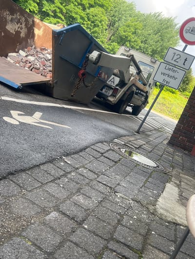 An outdoor, low-angle, eye-level shot shows a construction debris dumpster in the city of Bochum, Germany. The dumpster is dark grey and blue, filled with broken bricks and debris, and is mounted on the back of a white truck. To the right of the dumpster are two road signs. The upper sign is a red circle with a truck inside, indicating a weight limit. The lower sign is rectangular and contains text and a number. The text reads "Liefer- verkehr frei" and below that "12 t". This signifies that delivery traffic is free up to 12 tons. The dumpster is parked on asphalt, and a pedestrian symbol is painted on the road. The sidewalk is paved with grey cobblestones. A manhole cover is visible on the sidewalk, reflecting the sky. In the background, there are green trees and a brick building. The lighting suggests it is daytime, with overcast skies.