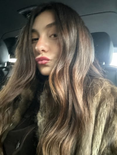 The image displays a close-up, frontal selfie of a young female individual situated within the interior of a vehicle. The subject possesses long, wavy, light brown hair with discernible highlights, flowing over her shoulders and part of her chest. Her lips are pursed in a pouting expression, accentuated by dark pink lipstick. She is attired in a dark, presumably black, turtleneck garment beneath a substantial brown fur coat or jacket. The immediate background reveals the dark upholstery and headrests of car seats, consistent with a vehicle's interior. No specific external landmarks or identifying features are present within the frame to establish a precise location within the city of Saint-Cyr-l'École, France; the scene is confined to the generic compartment of a private automobile.