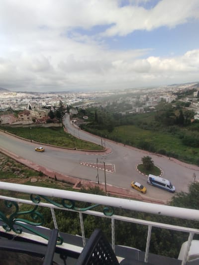 The image presents an elevated view of Tétouan, Morocco, likely captured from a balcony. The foreground features a white and green railing with a glimpse of outdoor furniture. The midground consists of a winding road with a roundabout, where two yellow taxis and a white and blue van are visible. The background reveals the urban sprawl of Tétouan under a cloudy sky. Vegetation, including grass and trees, punctuates the landscape.