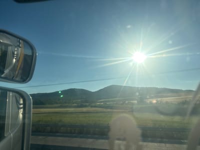 The image depicts a view from inside a vehicle, looking outwards through the windshield and side mirrors. The sun is intensely bright, high in a clear blue sky, creating lens flares and a strong glare across the scene. In the foreground, the chrome-trimmed housing of a side-view mirror is visible on the left, reflecting the side of the vehicle, which appears to be yellow. Below the mirror, another, smaller side-view mirror is partially visible. The view through the windshield shows a rural landscape with rolling hills covered in vegetation. The hills are green at their base and transition to drier, lighter-colored vegetation further up. A thin line of power lines is visible against the sky in the middle distance. The foreground outside the vehicle shows a grassy area, possibly a roadside verge, and a section of road or pavement. There are no people visible in the image. The scene appears to be set in a rural area during daytime, under a bright sun. The mention of Λύρα, Greece, suggests a potential geographical context, though the visual elements themselves do not definitively confirm or provide specific details about this location.