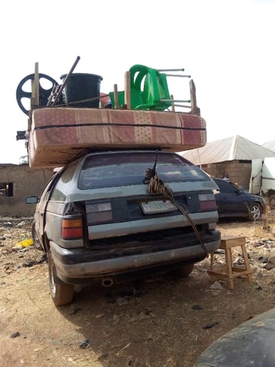 A faded gray wagon is overloaded with a mattress, wheel, bucket, and green chair strapped to its roof. The car is parked on a dirt ground with trash scattered around. A license plate is barely visible on the back. A wooden stool sits to the right of the car. Other vehicles and structures, including a tent, are visible in the background. The overall impression is one of poverty and precariousness.