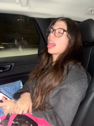 A woman with long brown hair and glasses is sticking her tongue out. She is wearing a gray sweater and appears to be leaning against the interior of a car, possibly in a backseat. Her eyes are closed, and her head is tilted to the side. A pink blanket or garment and a patterned blue tote bag are visible in the lower right corner of the frame. A phone is visible near her hands. The car's interior includes a black door panel and window.