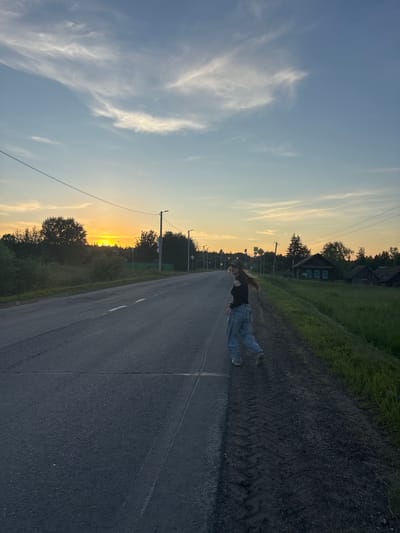 A young woman with long, light brown hair is captured mid-stride on the right side of a paved road in a rural setting, likely near Okulovka, Russia. She is wearing a dark off-the-shoulder top and loose-fitting light blue jeans, her body angled away from the camera as she looks over her left shoulder. Her right leg is extended forward, and her left leg is bent at the knee, suggesting movement. Her hair is flowing behind her, indicating a brisk pace or a breeze.
The road, asphalt, stretches into the distance, appearing somewhat worn with visible cracks and lane markings. To the right of the road is a gravel shoulder with distinct tire tracks, and beyond that, a vibrant green grassy field. Scattered throughout the field and along the horizon are several rustic wooden houses with dark roofs and small, blue-paned windows. Tall trees and dense foliage line the left side of the road, obscuring the view beyond.
The sky is a gradient of pale blue at the top, transitioning to a soft yellow and orange near the horizon, indicative of a sunset or sunrise. Wispy, white clouds are scattered across the upper sky. Several utility poles with visible wires are positioned along the left side of the road, and streetlights are intermittently placed further down the road. The overall impression is one of a serene, yet dynamic, rural landscape at dusk.