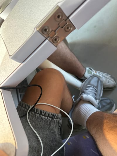 The image shows a close-up view of two pairs of legs from a seated perspective, possibly on public transportation or in a waiting area. The subjects are wearing athletic shoes and casual clothing. One person is wearing grey and white sneakers with white socks, and denim shorts. The other person's legs are partially visible, wearing similar grey and white sneakers and white socks. One of the individuals has a phone charging cable plugged in, with both black and white wires visible. The setting appears to be indoors with muted lighting. One leg is positioned over the other. The overall impression is one of casualness and everyday activity. There is no visible text or information that suggests a specific time of day, weather, or strong emotions. The location context of Narbonne, France, doesn't offer any specific visual cues within the image itself. The scene is dominated by the lower bodies of the individuals, with a focus on their footwear and attire.