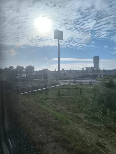 The image depicts a view from a moving vehicle, likely a train, looking out towards a suburban or industrial area on a bright, cloudy day. The sun is visible in the upper left portion of the sky, partially obscured by dense, horizontally layered clouds. In the mid-ground, a tall, slender pole with a square structure at the top is prominent, possibly an advertisement display or a communication tower. To the right of this structure, a water tower with a distinctive cylindrical base and a cubic reservoir on top stands against the skyline.

The foreground consists of a grassy embankment with some gravel or track material visible on the left. A black metal fence runs horizontally across the scene, delineating a path or boundary. Beyond the fence, the terrain is covered in lush green grass, with some taller vegetation on the right.

In the middle distance, there is a road or intersection with traffic lights and street lamps. Two individuals are seen on bicycles on this road, one on the left and one on the right. In the far background, several buildings are visible, including what appears to be a McDonald's restaurant with its distinctive golden arches logo, and other low-rise structures. Trees are present in the mid-ground, suggesting a developed area. The overall impression is a typical roadside view in the Netherlands, with infrastructure and commercial establishments.
