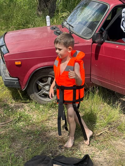 A young boy, likely pre-teen, is lying on his back on the ground next to a red car. He is wearing an orange life jacket with black straps. His legs are extended and his feet are dirty. The car is parked on an unpaved surface with grass and dirt. The boy's head is turned towards the car, and his facial expression is not clearly discernible. A portion of the car's door, window, side mirror, and wheel are visible. The scene suggests an outdoor setting, possibly a rural or recreational area. The lighting indicates it is daytime, and the weather appears to be fair. The context provided places this scene in Pravda, Russia. There is text visible on a can lying on the ground near the car, which reads "100". The overall mood of the image is casual and appears to capture a moment of rest or preparation before or after an outdoor activity.