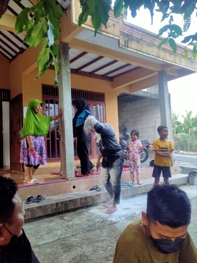 The image captures an outdoor event in Celep, Indonesia, taking place on a concrete ground area adjacent to a residential building. In the foreground, two adult males are partially visible; the male on the left is seen from the side, while the male on the right wears a black face mask and an olive-green shirt, looking downwards.
Centrally, an adult individual with short dark hair, wearing a dark long-sleeved top and dark jeans, is heavily covered in white powder and appears wet. This individual is bending forward, standing barefoot within a significant spread of white powder on the ground. Multiple pairs of footwear, including black slippers, are visible on the elevated porch steps.
On the elevated porch of the house, an adult female in a bright green hijab and a purple patterned skirt stands barefoot, smiling and leaning against a light gray concrete pillar. Behind the powdered individual, another adult female wearing a black hijab and dark attire stands barefoot on a red patterned mat on the porch.
Two children observe the scene from the ground level on the right. A young girl in pink floral pajamas stands behind the powdered individual, near a dark motorcycle parked on the unpaved area. A young boy in a yellow striped t-shirt and dark shorts stands closer to the powdered individual, smiling towards the left. A gray bucket is visible to the far right.
The residential building features a light orange facade with brown wooden windows and doors. An extended roof, supported by the central concrete pillar, covers the porch. Green leaves from an overhead tree partially obscure the upper left corner of the frame. The background reveals additional residential structures, sparse vegetation, including banana plants, and a dirt road or path. The scene is illuminated by daylight.