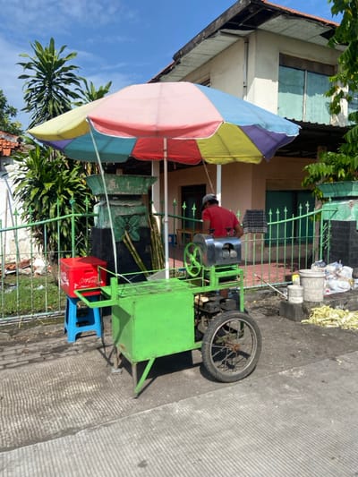 A man wearing a red shirt and a baseball cap is standing next to a green cart with a red cooler next to it. He is standing in front of the cart, facing away from the camera. There is a large, multi-colored umbrella above the cart and man. The cart appears to be a street vendor cart with a large, metal drum-like object attached to the cart. The street vendor cart appears to be selling sugar cane juice. There is a metal fence behind the cart, and a concrete building to the right of the fence. The scene is located in Kecamatan Tulangan, Indonesia. The coordinates are -7.482 latitude, 112.648 longitude.