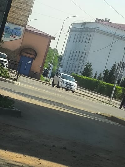 This image captures a street scene in Abay, Kazakhstan, bathed in the bright, clear light of midday. The foreground is dominated by a dusty, unpaved area leading to a paved road. On the left side of the road, a small brick building with a brown roof is visible, featuring a blue door and a sign with blue lettering that reads "Pan" in a stylized font, followed by the word "кафе" (cafe) below it in a smaller, plainer script. This suggests a local cafe is present.
Further down the road, a white car is driving, its headlights on. Beyond the road, a large, white, multi-story building with a red roof stands prominently, characterized by its repetitive window pattern. The surrounding area is lush with green trees and bushes, indicating a pleasant, possibly warm weather day. In the far right of the image, a person in dark clothing is walking away from the camera. The overall impression is that of a typical urban or suburban street in a Central Asian town, with a mix of older and newer architecture, and daily life unfolding under a bright sky. The scene is devoid of any obvious signs of urgency or distress, suggesting a calm, ordinary day.