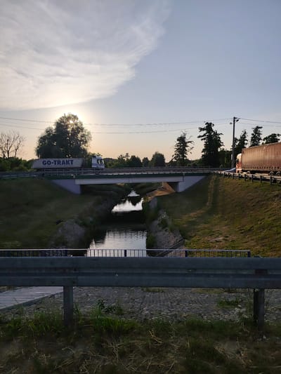 The image shows a bridge over a canal in Stryków, Poland. The sun is setting, casting a warm glow over the scene. A white truck with "GO-TRAKT" written on its side is driving across the bridge. To the right, a red truck with "Palifor Logistics" written on it is also visible. The canal below is calm and reflects the sky and the bridge. Green grass and trees line the banks of the canal and surround the bridge. A metal guardrail is in the foreground, obscuring some of the view of the canal and the bridge. Electrical wires are visible stretching across the sky.