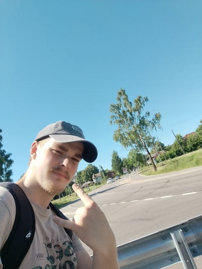 The media file is a selfie of a young man standing outdoors. He is wearing a grey baseball cap, a light-colored t-shirt with a graphic print, and a black backpack. He has light-colored hair and a mustache, and appears to be in his late teens or early twenties. He is making a "rock on" hand gesture with his right hand, pointing towards the viewer with his index finger. The background shows a rural road intersection with some trees and houses visible. The sky is clear and blue, indicating a sunny day. The time of day appears to be afternoon, given the lighting. The man's expression is neutral, but the gesture and the selfie format suggest a casual and possibly playful mood.
The scene is set in Dižakmens, Latvia. The road appears to be paved, with painted white lines indicating lanes. There are also some tire marks visible on the road surface. A metal guardrail is present in the foreground, suggesting this may be near a road or highway. In the background, there are several trees, a tall utility pole with wires, and some residential buildings. A traffic sign is also visible in the distance. The overall setting is one of a typical rural environment in Latvia.
The t-shirt has some visible text that appears to be printed in a distressed font, but it is mostly obscured by the man's arm and the image quality. Some of the letters are visible, like "WRM" and parts of other words in a circular pattern. The baseball cap also has a logo or design on it, but it is not clearly discernible.