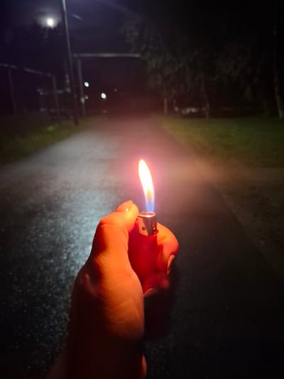 A first-person perspective shot shows a hand holding a red lighter with a flame burning at night. The lighter is illuminated by the flame, casting an orange glow on the hand. The background is a dark, blurred path, possibly a road or walkway, with some greenery on the sides. There are some out-of-focus lights and what appears to be the moon in the upper left corner. The scene is likely outdoors in a dimly lit area. The location in the city of Novlyanka, Russia is not discernible from the image itself.