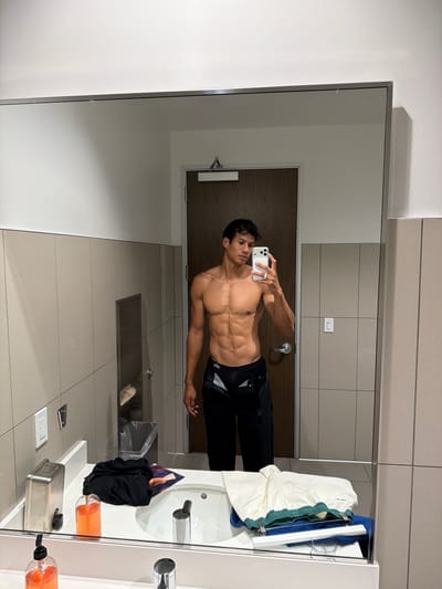 A shirtless, muscular man is taking a selfie in what appears to be a public restroom. He is wearing black athletic pants. The restroom has light-colored tile walls and floors. A sink with soap dispensers and clothes are visible to the right. A door and other restroom fixtures are visible in the background. The location is in the city of Torrance, United States of America.
