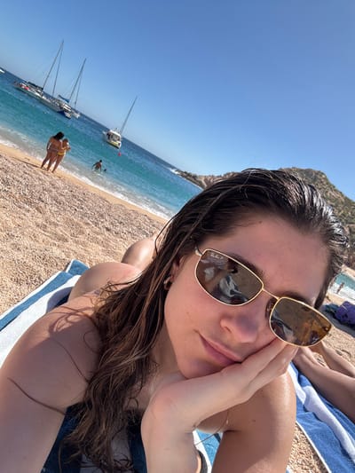 A selfie-style photo captures a person relaxing on a sunny beach. The main subject, presumably female, is positioned in the foreground, filling the right side of the frame. She has long, dark brown hair that appears wet, suggesting she may have recently been in the water. She is wearing gold-rimmed sunglasses, one of which clearly displays "Ray-Ban P" on the lens. The sunglasses reflect an indistinct shape, possibly another person or object nearby. Her left hand is resting against her cheek, and she is looking directly at the camera with a relaxed expression. Her skin appears tanned and possibly glistens from water or sunscreen. She is lying on a blue and white striped towel, with parts of her bare shoulder and arm visible.
In the midground, to the left of the main subject, a pebbly or sandy beach stretches alongside clear, turquoise blue water. Several individuals are visible enjoying the beach. Two people are standing together on the beach in the lower left, appearing to be wearing swimsuits. Further out in the water, another person is partially submerged. Several white sailboats are anchored in the calm water, with at least two clearly visible multi-masted boats.
The background consists of a clear, bright blue sky, indicating a sunny day. A rocky landmass or mountain rises from the water on the far right, suggesting a coastal or island setting. The overall impression is one of a vacation or leisure activity at a scenic beach location. The image is taken at a slight tilt, characteristic of a casual selfie.