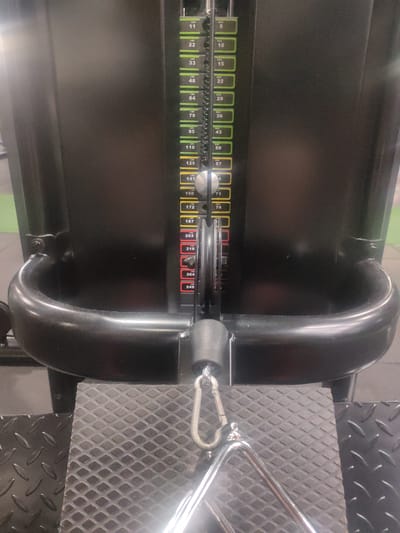 An indoor, close-up, eye-level shot shows the weight stack and cable pulley of a black gym machine. The weight stack consists of multiple rectangular plates stacked vertically, with labels indicating weights in both pounds (LBS) and kilograms (KGS). The current selected weight is 202 LBS / 92 KGS, as indicated by the red indicator on the stack. A silver pin is inserted into the stack to select this weight. A black cable emerges from the top of the stack and runs through a pulley mechanism, connecting to a carabiner and a silver cable attachment, likely a triangle bar, at the bottom of the frame. The machine is positioned on a textured black platform. The background is a dark, matte black surface. The scene suggests a gym environment, with no people visible. The image lacks any contextual elements to ascertain its location in Figueres, Spain.