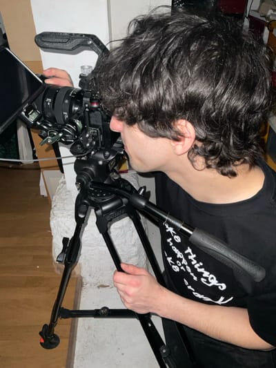 A person with dark, curly hair is shown from the side, leaning over a camera mounted on a black tripod. The camera has a lens attached and various accessories. The person is wearing a black t-shirt with white text. The tripod legs are extended and rest on a floor with mixed textures. The background shows a glimpse of a white-covered surface and some darker objects. The image is cropped tightly around the person and the camera equipment.