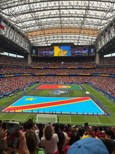 This is a high-angle, wide shot of a soccer stadium during a pre-game ceremony. The main subjects are a large contingent of people, including players lining up on a soccer field and a massive crowd filling the stadium seats. The field itself is dominated by a large replica of the flag of the Democratic Republic of Congo, with its distinct blue, red, and yellow colors, and a yellow star. Players, dressed in matching blue uniforms, stand at attention along the edges of the flag. The stadium is vast and appears to be covered by a large, intricate metal roof structure.
The event taking place is likely the pre-game national anthem or introductions, as suggested by the formal lineup of players and the presence of the flag. The atmosphere seems anticipatory, with the large crowd indicating a significant event. The lighting suggests it is daytime, and the setting is an indoor stadium, likely in Houston, Texas, given the location context.
Notable details include the large video screens visible at one end of the stadium, displaying images and text. One screen shows a close-up of what appears to be a soccer team and text in French: "L'HYMNE SACRÉ DE VOTRE SOLIDARITÉ." Below this, another screen displays "FIFA WORLD CUP..." and the number "5" with the year "2026". Another smaller screen shows text in what appears to be Latin script, possibly listing country names, including "CONGO". The crowd in the stands is densely packed, with many individuals wearing red and white clothing. The overall impression is of a formal and energetic sporting event.