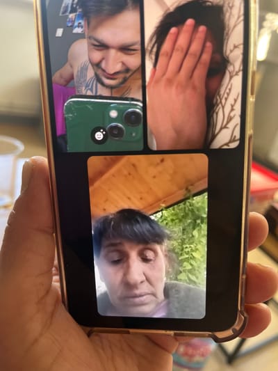A hand holds a smartphone displaying a video call with three participants. One participant is in bed, covering their face with a hand. Another participant is outdoors, near greenery. The third participant is indoors and has tattoos on their upper body. The location is unknown, as it cannot be determined from this image.