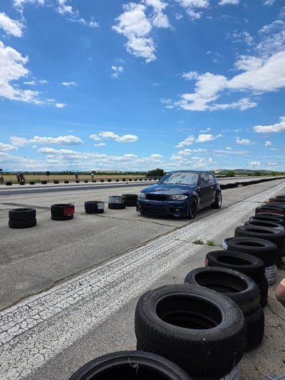 The image depicts a drag strip or race track under a bright, partly cloudy blue sky. In the foreground, a blue BMW hatchback is parked on the asphalt. The track is lined with stacks of tires, some painted black and red, serving as a visual barrier. The surface of the track shows wear and cracking in places. In the background, several people are visible standing near the edge of the track, and a dry, field-like landscape stretches to the horizon, dotted with distant trees and rolling hills. The overall scene suggests an outdoor event or practice session at a racing facility. The location of Akatsievo, Bulgaria is not discernable from the image content itself.