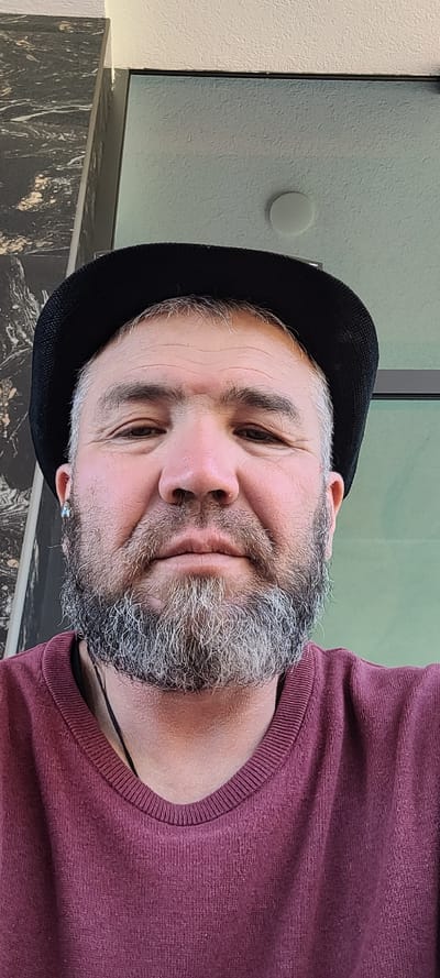 A close-up, head-and-shoulders shot, taken from a low angle, features a middle-aged Caucasian male wearing a black brimmed hat and a maroon crewneck sweater. The man has a salt-and-pepper beard and mustache, with visible wrinkles on his forehead and around his eyes. He is looking directly at the camera. An earring is visible in his left ear. He is wearing earbuds. The background consists of a marble-patterned wall on the left and a blurred green surface on the right, suggesting an indoor or covered outdoor setting. A white circular light fixture is visible in the upper right portion of the background. The lighting appears to be artificial, casting a soft glow on the subject's face. The image is likely a selfie.
