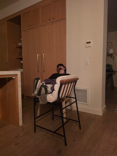 This media file captures an indoor scene, likely a residential kitchen or dining area in Coronado, United States of America. The image is oriented sideways, but conceptually, it shows a person resting or sleeping in a tall, bar-stool style chair.

The subject is an individual, seen from the side, with their head tilted back and mouth slightly open, suggesting they are deeply asleep or in a state of profound relaxation. They are wearing a dark cap and a dark long-sleeved top. Their feet, encased in white and grey sneakers with dark soles, are propped up on the chair's rungs. The chair itself has a wooden seat and backrest, complemented by a black metal frame. A striped cushion and a soft, light-colored, possibly terry cloth, blanket or towel are draped over the chair's back.

The setting includes light-colored wooden cabinetry or built-in furniture with brass-colored handles and small knobs, extending across the left side of the frame. The wall behind the chair and cabinets is plain white, featuring a white rectangular vent, a white thermostat or control panel, and a white light switch. The floor is made of light brown wooden planks. In the background (upper right when viewed upright), a wall-mounted lamp and a small potted plant are partially visible. A thin white cable lies on the wooden floor in the foreground, leading towards a counter-like surface.

The lighting appears to be artificial, suggesting the photo was taken indoors during the evening or at night. The scene conveys a quiet, personal moment of rest. No discernible text is visible on the wall fixtures or other objects.