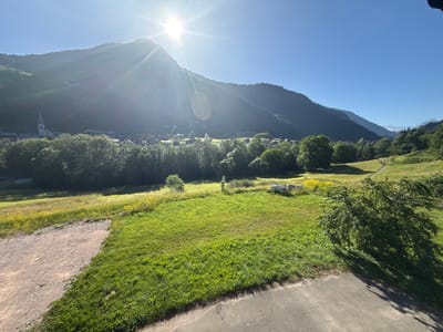 A bright, sunny day in Saint-Jean-d'Aulps, France, illuminates a scenic vista. The sun, positioned in the upper left quadrant, shines intensely, casting lens flare and rays across the sky and down the mountainside. A lush green meadow dominates the foreground, transitioning into a mix of green grass and yellow wildflowers further back. A gravel path cuts diagonally across the bottom left of the frame, meeting the paved surface in the lower right. In the mid-ground, a cluster of trees separates the meadow from a village nestled at the base of a steep, tree-covered mountain. The village features buildings, including a prominent white church with a tall steeple. Beyond the village, the mountain rises sharply, its slopes covered in dark green trees, with the sun's light creating a bright halo effect at its peak. The sky is a clear, vibrant blue.