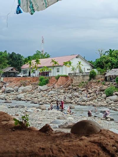 This detailed image captures a daytime scene at a rocky riverbank in Meurah, Indonesia, under an overcast sky. In the immediate foreground, dark brown soil and a small green plant are visible, with a large, rounded dark rock partially obscured. The midground features a wide, shallow riverbed densely covered with numerous grey boulders and smaller rocks, through which water flows. Several individuals are present in and around the river; some are wading in the water, others sit on the rocks. People are dressed in casual attire, with several women wearing hijabs.
On the riverbank, an eroded dirt slope transitions to a more elevated area. To the left, a simple, open-sided wooden structure with a corrugated roof stands next to a white pickup truck. Behind this, a prominent white building with a red-orange gabled roof is situated on a raised, concrete foundation. Palm trees are planted in front of this building, which features multiple windows and a porch area. The bank below the building shows significant erosion, exposing reddish soil and scattered debris. To the right of the white building, another smaller, thatched-roof structure is visible, with people gathered inside or nearby.
Beyond the buildings and structures, dense green foliage and trees line the horizon, with mountains faintly visible in the far distance. A tall, white communication tower with multiple antennas rises above the treeline on the left side of the background. At the very top edge of the image, partially obscured, is a piece of light-colored fabric or tarp with green markings, from which a small blue plastic bag hangs.