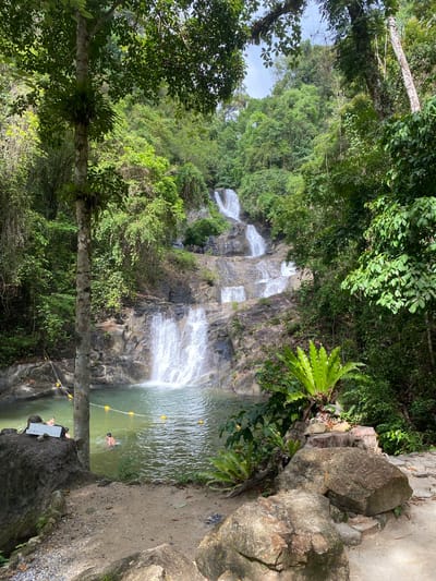 The image captures a multi-tiered waterfall cascading down a rocky cliff face surrounded by lush green vegetation. The location is Tambon Thai Mueang, Thailand. The waterfall empties into a pool where a person is swimming. A safety rope marked with yellow buoys spans the width of the pool. A tree trunk stands vertically in the foreground, partially obscuring the view. On the right side of the image, a rocky outcrop with a concrete path runs along the edge of the pool. Two people are present near the rocky outcrop and the safety ropes.