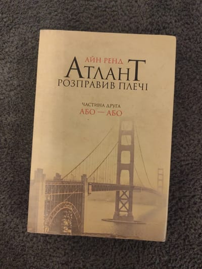 The image shows a book with a beige cover, featuring a sepia-toned photograph of the Golden Gate Bridge. The title of the book, in Cyrillic script, is visible at the top. The first line reads "АЙН РЕНД", followed by a large, stylized "АТЛАНТ". Below this, the title continues "РОЗПРАВИВ ПЛЕЧІ". Further down, smaller text reads "ЧАСТИНА ДРУГА" and "АБО — АБО". The book is lying flat on a dark, textured surface, likely carpet. There are no people or animals visible in the image. There is no information in the image that would allow for determining its location in the city of Schwäbisch Gmünd, Germany.