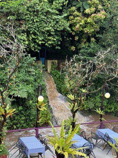 The image shows an outdoor seating area with tables covered in blue and white checkered tablecloths, set up near a brown river. A waterfall is visible in the background, cascading down a rocky embankment covered in lush green vegetation. Two bare trees with small orange lights are in the foreground, and ferns and other plants are interspersed throughout the scene. A purple railing separates the seating area from the river. The area appears to be a restaurant or cafe in Sikeben, Indonesia, offering scenic views of the natural landscape. There are no people visible in the image.