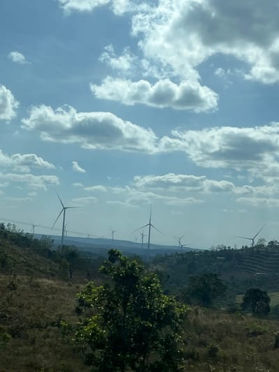 A scenic view features a multitude of wind turbines standing against a backdrop of rolling hills, partially obscured by vegetation. The sky is filled with fluffy, white cumulus clouds floating in a blue expanse. The horizon line is hazy, creating a sense of depth and distance. A few green trees are on the right of the landscape. The composition is vertically oriented.