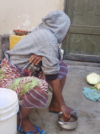 A person in a gray hoodie and colorful patterned skirt sits near a white bucket. They are mixing something in a silver bowl with their hand. A yellow container and a dish of red peppers are behind them. Next to them, a halved cabbage and a light blue cloth sit on the ground near a green door. They wear blue flip-flops. The scene appears to be outdoors, possibly in a courtyard or backyard.
