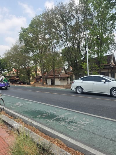 The image shows a street scene in the Historic City of Ayutthaya, Thailand, with a white car parked partially on a green bike lane. The bike lane has faded white lettering indicating its purpose. A street light stands near the car. The background features traditional Thai houses with tiled roofs, partially obscured by large trees. The sky is visible through the branches. A partial view of a person and a purple vehicle is at the bottom of the frame. Vegetation grows along the edge of the bike lane.