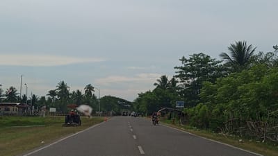 The image depicts a rural road in Glokaron, Indonesia, with lush green vegetation and palm trees lining both sides. The sky is overcast with soft clouds.
On the left side of the road, there is a grassy area with a small building and an Indonesian flag flying from a pole. A tractor is visible in the foreground, with a person operating it, and a white mist is being sprayed from it, likely for irrigation or pest control. Several other structures and what appear to be temporary barriers are also present in this area.
The road itself is paved and has a dashed white line marking the center. Several vehicles are on the road, including motorcycles with riders. On the right side of the road, more dense green foliage and trees are visible, with a small, open-sided shelter.
The overall impression is one of a working rural landscape on a cloudy day.