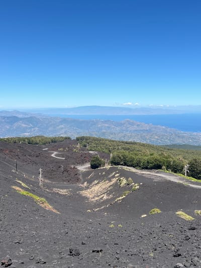 The image captures a panoramic view of Mount Etna, a volcano located near Linguaglossa, Italy. The foreground is dominated by dark, volcanic rock, indicative of a recent eruption or solidified lava flow. A winding dirt road snakes through this barren landscape, leading the eye towards a lush, green forest that carpets the middle ground. Beyond the forest, the terrain becomes mountainous and rugged, with layers of hills fading into the distance. In the far background, a vast expanse of blue water, likely the Ionian Sea, stretches to the horizon, with a distant coastline visible under a clear, bright blue sky. The overall scene is one of raw, natural beauty, showcasing the dramatic landscape of volcanic activity juxtaposed with the resilience of nature.