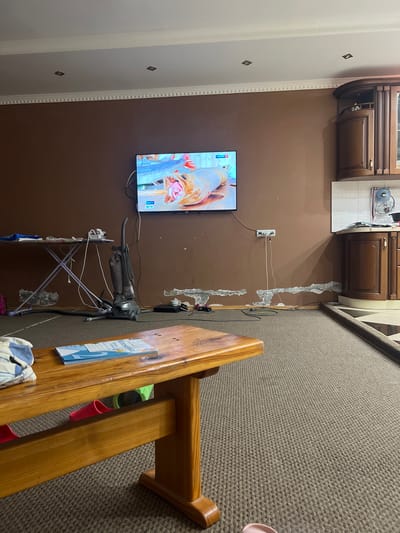 The image depicts an indoor scene, likely a home in Vodnik, Russia. A large television is mounted on a brown wall, displaying a close-up of fish being prepared, with the "РОССИЯ 1" logo visible in the bottom left corner. To the right of the TV, an ironing board with a vacuum cleaner attached to it stands against the wall. Various cables are connected to an electrical outlet above the ironing board.
Above the television and ironing board, dark brown kitchen cabinets are visible, featuring metallic handles. The wall itself is painted a muted brown, and to the left, a white wall with decorative molding and diamond-shaped accents suggests a transition to another room.
The scene continues to the right with a textured, grey carpet covering the floor. A wooden staircase or furniture piece with a light brown finish is prominent in the foreground, showcasing its grain and construction. A blue booklet with Russian text, possibly a school notebook, is attached to the wooden structure. A pair of pink, rubber-like objects and a green object are placed within or near the wooden furniture. The overall impression is of a lived-in space, with everyday items present. There are no people visible in the image.